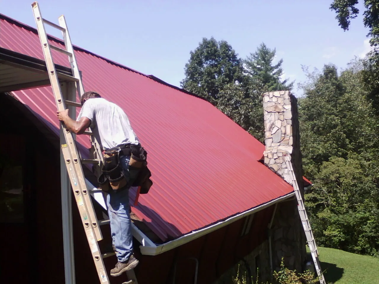 Roofer on a ladder beside a red metal roof and stone chimney — Soffit & Fascia Repair by Roofing Brothers in Vidor